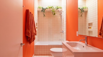 Baño con paredes naranjas, toallas colgadas, inodoro blanco, lavabo con grifo, espejo y bañera azulejada con plantas en un estante superior.