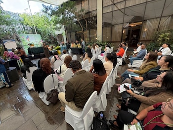 Personas sentadas en sillas blancas asisten a una exposición rodeada de plantas y arreglos florales