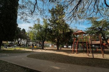 Campo de flores amarillas y hierba verde. Una fila de árboles oscuros y un edificio largo. Cielo con nubes en tonos anaranjados, amarillos y azules