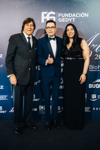 Dos hombres con traje y una mujer con vestido largo negro posan frente a un fondo azul con logotipos de la Fundación Gedyt