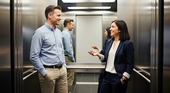 Hombre y mujer conversan y sonríen dentro de un ascensor con paredes metálicas. Él tiene las manos en los bolsillos y ella gesticula con una mano.