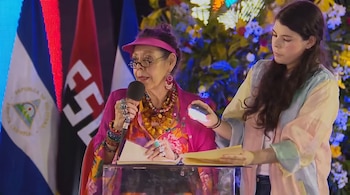 Una mujer con visera rosa y micrófono, y otra mujer con una luz y papeles en un atril. Al fondo se ven banderas de Nicaragua y del FSLN con decoraciones.