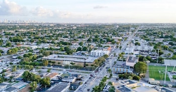 Vista aérea de Hialeah con casas, árboles, calles con tráfico y edificios, y el horizonte de Miami al fondo bajo un cielo parcialmente nublado