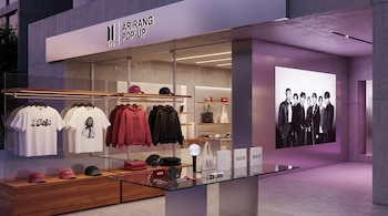 Fachada moderna de la Pop-Up Store 'BTS ARIRANG POP-UP'. Exhibidor con camisetas, sudaderas, gorras, lightstick y álbumes; pantalla digital de BTS y luz morada.