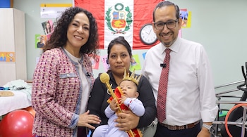 Una madre sostiene a un bebé coronado entre un hombre con gafas y una mujer con chaqueta estampada ante la bandera de Perú