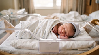 Un bebé recién nacido con gorro a rayas duerme envuelto en una manta blanca dentro de una cuna de plástico transparente.