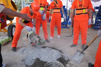 Grupo de trabajadores con uniformes naranjas y cascos, uno vierte cemento sobre arena en el suelo. Hay una pala, una hormigonera y sacos de cemento