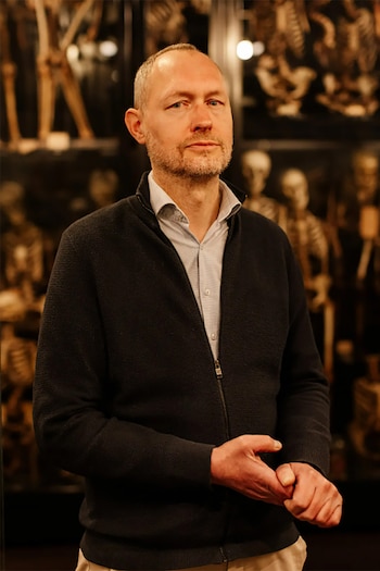 Retrato de Laurens de Rooy, un hombre de mediana edad con barba, suéter oscuro y camisa clara. El fondo borroso muestra vitrinas con esqueletos