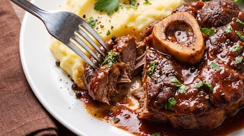 Un tenedor corta un trozo de osobuco con hueso sobre puré de papas cremoso, bañado en salsa oscura, decorado con granos de pimienta y perejil fresco.