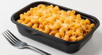 Macarrones con queso servidos en un recipiente negro para comida preparada. Un tenedor metálico descansa al lado sobre un fondo blanco.