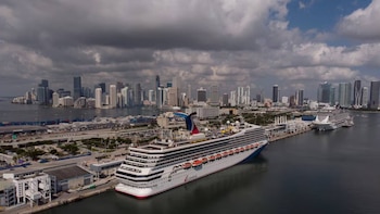 Vista aérea de dos grandes cruceros atracados en PortMiami, con el horizonte de rascacielos de Miami al fondo y un cielo parcialmente nublado