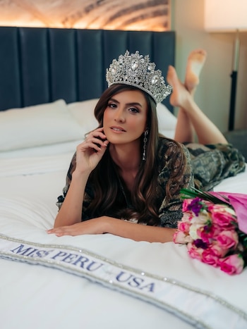Una mujer joven con cabello castaño y una corona de diamantes, vistiendo una bata brillante, acostada en una cama blanca junto a un ramo de rosas y una banda de Miss Perú USA