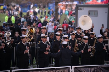 Músicos uniformados con gorras blancas tocan clarinetes, saxofones y una tuba frente a un director y espectadores