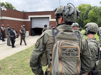 Vista posterior de agentes de seguridad en uniforme táctico y cascos, uno con mochila que dice "SBI AGENT BOMB TECH", frente a un edificio de ladrillo con portón abierto