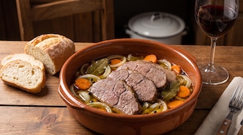 Cazuela de barro con rebanadas de carne, zanahoria, cebolla y hojas de laurel, junto a rodajas de pan y una copa de vino tinto.
