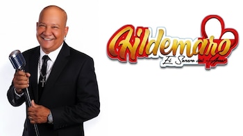 Hombre calvo, traje negro, corbata negra, cadena con cruz, reloj, anillo, sonriendo, sosteniendo un micrófono plateado, logo Hildemaro