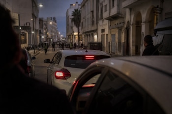 Un automóvil blanco con las luces de freno encendidas permanece detenido en una calle flanqueada por edificios al anochecer.