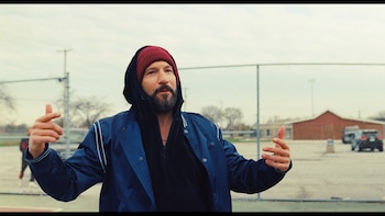 Un hombre barbudo, con gorro rojo y sudadera negra bajo una chaqueta azul, gesticula con ambas manos al aire libre, con una valla metálica y edificios al fondo