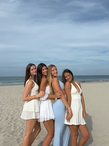Cuatro mujeres jóvenes posan sonrientes abrazadas en una playa de arena con el mar y el cielo azul al fondo