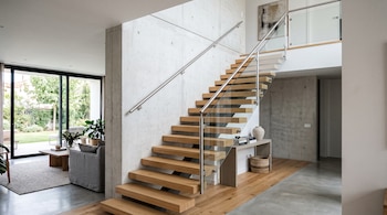 Escalera recta con peldaños de madera y barandilla de vidrio junto a un muro de hormigón, con una sala de estar al fondo.