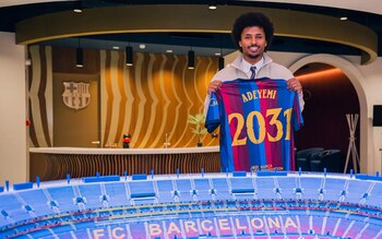 Hombre sonriente con camiseta del FC Barcelona que dice "ADEYEMI 2031", frente a una maqueta de un estadio y el escudo del club