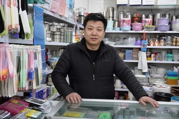Un hombre de pie detrás de un mostrador de cristal en una tienda rodeado de estanterías con utensilios y productos de plástico.