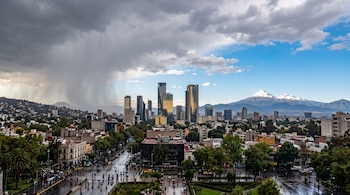Vista panorámica de una ciudad con rascacielos, calles mojadas, lluvia densa a la izquierda y montañas nevadas en el horizonte.