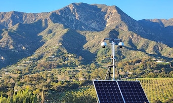 Una torre de vigilancia metálica con cámaras de seguridad y paneles solares se ubica frente a una montaña