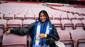 Una mujer con bufanda de Nicaragua, bandera, escudo, chaqueta negra y cámara Canon, sentada en gradas de un estadio con asientos rosas vacíos.
