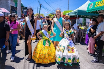 Hombre y mujer asiáticos sonriendo en primer plano, la mujer sostiene un letrero con forma de piña que dice "Feria de la Piña 2026 Santa María Ostuma"