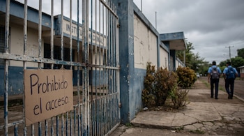 Reja azul y blanca de una escuela en Nicaragua con un cartel de "Prohibido el acceso"; dos estudiantes uniformados se alejan al fondo bajo un cielo gris nublado.