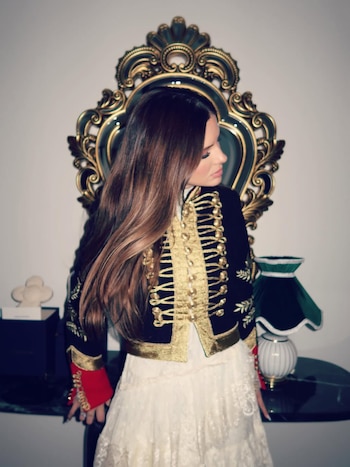China Suárez de perfil, mirando hacia la derecha, con una chaqueta negra y dorada estilo militar y una falda blanca de encaje, frente a un espejo ornamentado