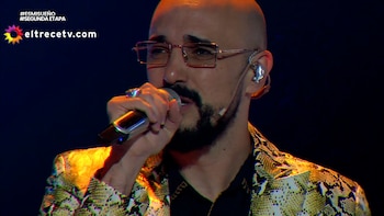 Primer plano de un hombre calvo con barba y gafas, cantando en un micrófono y vistiendo una chaqueta con estampado de serpiente durante una actuación en el escenario