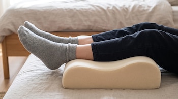 Primer plano de las piernas de una persona, con pantalones oscuros y calcetines grises, que descansan sobre una almohada ortopédica beige en una cama.