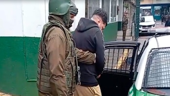 Un agente de seguridad con casco y chaleco táctico escolta a un hombre vestido de oscuro hacia un vehículo policial con rejas