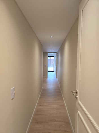 Pasillo estrecho con paredes beige claro, suelo de tablones de madera clara, luces empotradas en el techo, puerta blanca a la derecha y una salida al fondo