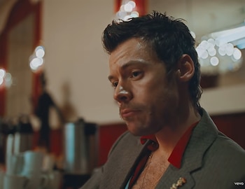 Primer plano de un hombre con el cabello corto, vestido con saco gris y cuello rojo, en un espacio interior con luces desenfocadas de fondo