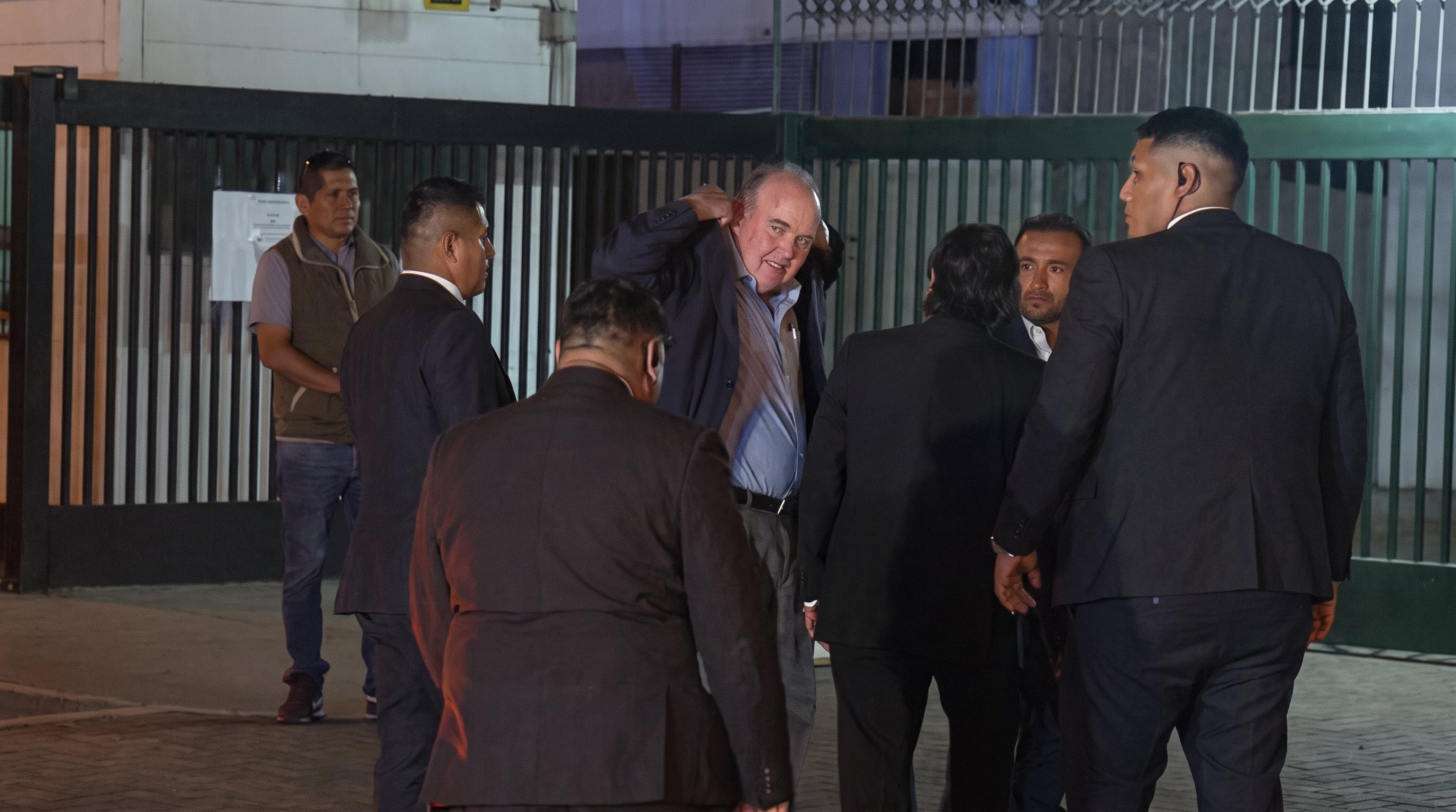 El político Rafael López Aliaga ajusta su saco mientras camina acompañado por personal de seguridad frente a una reja exterior. (Imagen Ilustrativa Infobae)