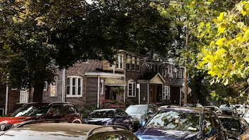 Vista frontal de una calle residencial con múltiples coches estacionados frente a casas de ladrillo rojo, rodeada de frondosos árboles verdes