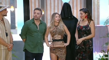 Cuatro personas y una figura cubierta de negro en un set. Un hombre con sombrero, un hombre con camisa verde, una mujer con vestido de leopardo, una mujer con vestido floral