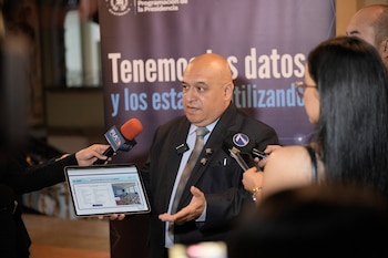 Hombre calvo con traje habla frente a micrófonos y muestra tableta con texto FICHAS DE PROYECTOS FINALIZADOS. Fondo con pancarta azul con texto