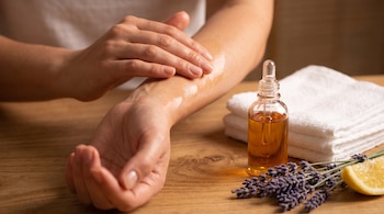 Unas manos frotan aceite en el antebrazo junto a un frasco gotero, toallas dobladas, flores de lavanda y una rodaja de limón sobre madera.
