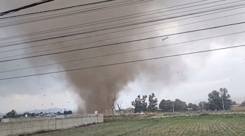 Un gran torbellino de polvo oscuro se eleva desde un campo verde bajo un cielo nublado, con cables eléctricos en la parte superior, una valla y árboles al fondo.