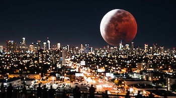 Una Luna grande parcialmente oscurecida con tonos rojizos sobre un horizonte urbano iluminado durante la noche.