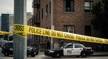 Cinta policial amarilla en primer plano; agentes tácticos de la DEA y LAPD frente a un edificio de ladrillos con vehículos de emergencia en California.