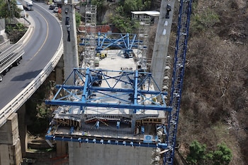 Vista aérea de un puente en construcción con pilares de hormigón y estructuras azules de soporte, junto a una carretera con vehículos y vegetación seca