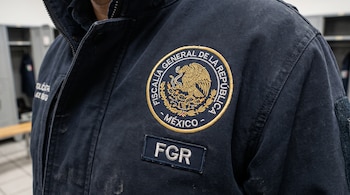 Primer plano del torso de una persona con chamarra azul marino que lleva un escudo bordado de la Fiscalía General de la República de México y un parche con las letras FGR.