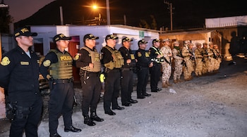 Una fila de agentes policiales vestidos de negro y soldados con uniformes de camuflaje permanecen de pie en una calle durante la noche.