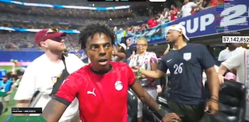 Un hombre de piel oscura con camiseta roja mira de frente. Detrás, otros hombres, una mujer y público en un estadio de fútbol. Pantalla indica 'ARG 3 EGY 2'