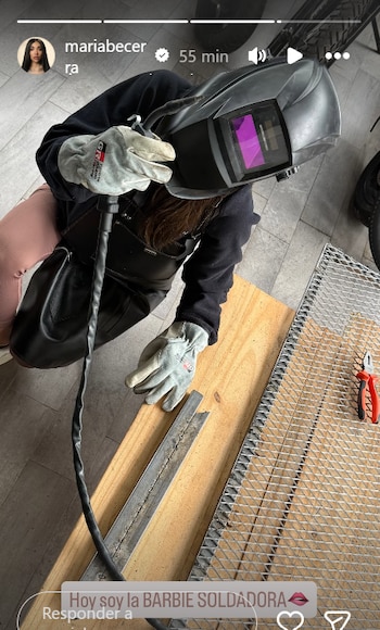 Vista superior de María Becerra soldando, con casco protector, guantes y delantal, trabajando en una reja metálica sobre una tabla de madera